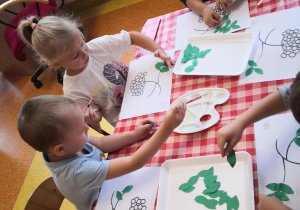 dzieci przy stoliku naklejają na kartkę listki jarzębiny we wskazanym miejscu